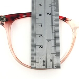 May include: Close-up of eyeglasses with a ruler measuring the frame. The glasses have a translucent pink frame with a dark red and brown patterned upper corner. The ruler shows a measurement of approximately 2 inches.