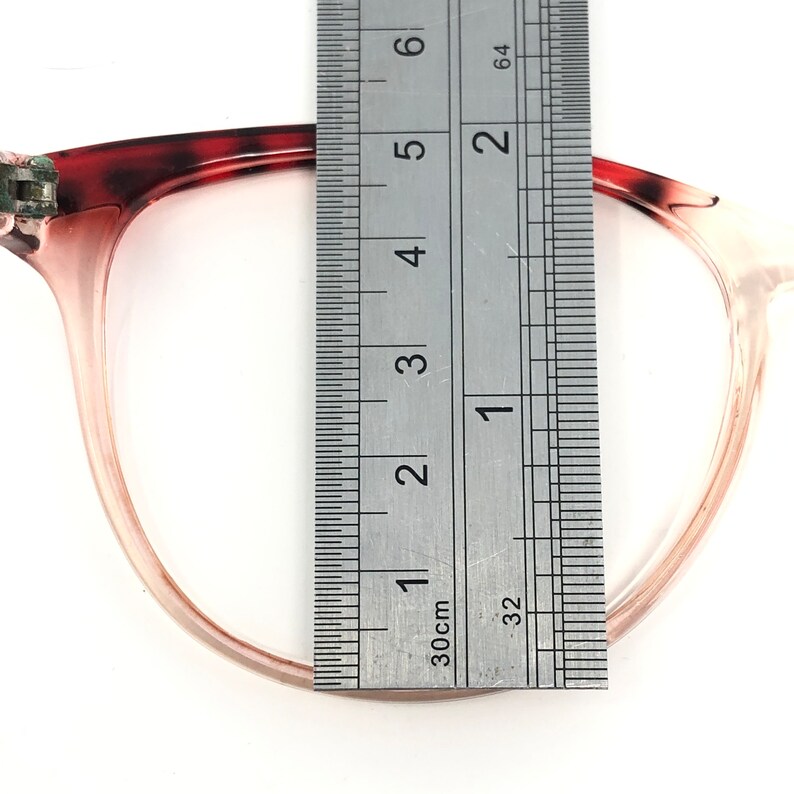 May include: Close-up of eyeglasses with a ruler measuring the frame. The glasses have a translucent pink frame with a dark red and brown patterned upper corner. The ruler shows a measurement of approximately 2 inches.