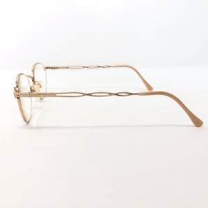 K&ouml;nnte beinhalten: Seitenansicht einer goldfarbenen Brille mit ovalen Gl&auml;sern. Die Fassungen haben ein filigranes, gedrehtes Design an den B&uuml;geln und gebogenen B&uuml;geln. Die Nasenpads sind hellbeige. Die Brille ist vor einem wei&szlig;en Hintergrund platziert.