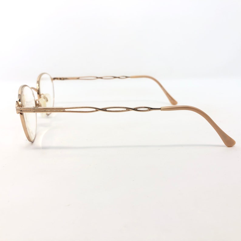 K&ouml;nnte beinhalten: Seitenansicht einer goldfarbenen Brille mit ovalen Gl&auml;sern. Die Fassungen haben ein filigranes, gedrehtes Design an den B&uuml;geln und gebogenen B&uuml;geln. Die Nasenpads sind hellbeige. Die Brille ist vor einem wei&szlig;en Hintergrund platziert.