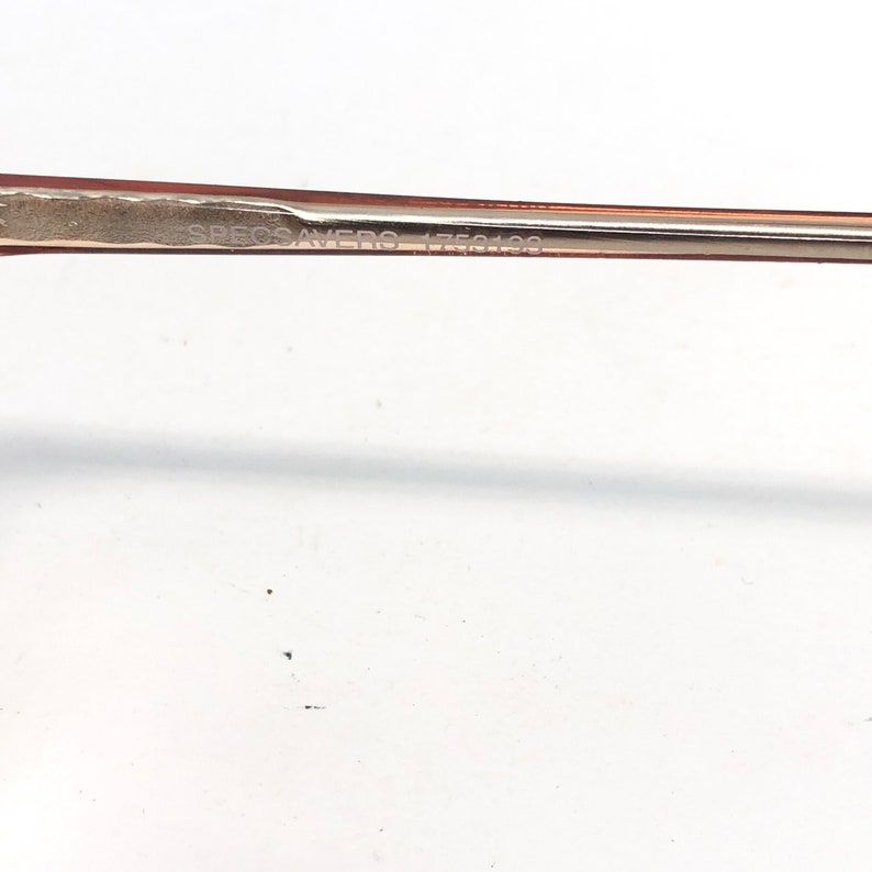 May include: Close-up of a pair of eyeglasses with a light brown frame. The text "SPECSAVERS 1750199" is printed on the frame. The background is white.