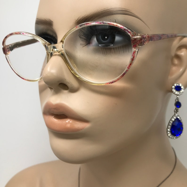 May include: Clear-framed eyeglasses with a floral pattern on the upper rims and temples. The glasses are on a mannequin. Dangling earrings with blue teardrop-shaped gems and clear stones are also visible.