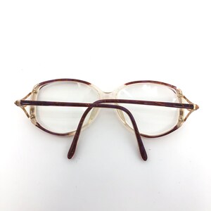 K&ouml;nnte beinhalten: Vintage Brille mit klaren Gl&auml;sern und einem hellbraunen und lila gemusterten Rahmen. Die Brille hat eine rechteckige Form mit einem d&uuml;nnen, goldfarbenen Metalldetail an den B&uuml;geln. Die B&uuml;gel sind dunkelbraun.
