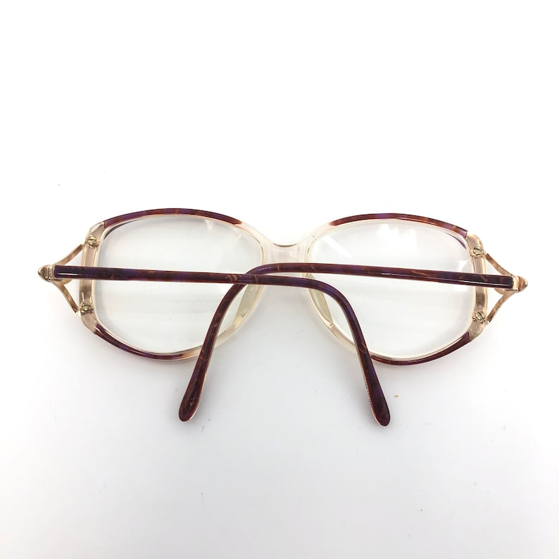 K&ouml;nnte beinhalten: Vintage Brille mit klaren Gl&auml;sern und einem hellbraunen und lila gemusterten Rahmen. Die Brille hat eine rechteckige Form mit einem d&uuml;nnen, goldfarbenen Metalldetail an den B&uuml;geln. Die B&uuml;gel sind dunkelbraun.