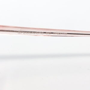 May include: Close-up of a portion of a pair of eyeglasses with a light pink frame. The text "SPECSAVERS SC 1988169" is printed on the frame. The background is plain white.