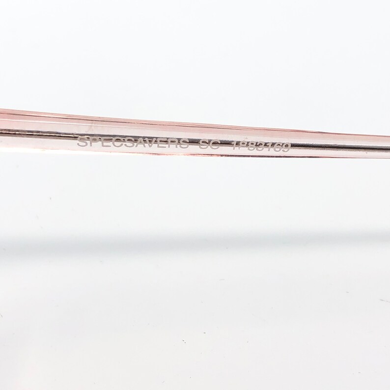 May include: Close-up of a portion of a pair of eyeglasses with a light pink frame. The text "SPECSAVERS SC 1988169" is printed on the frame. The background is plain white.