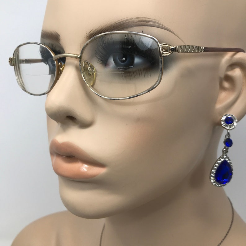 May include: A mannequin wears gold-framed spectacles with clear lenses. The glasses have a rectangular shape and patterned temples. Dangling earrings with blue, teardrop-shaped gems and silver detailing are also visible, adding a touch of elegance.
