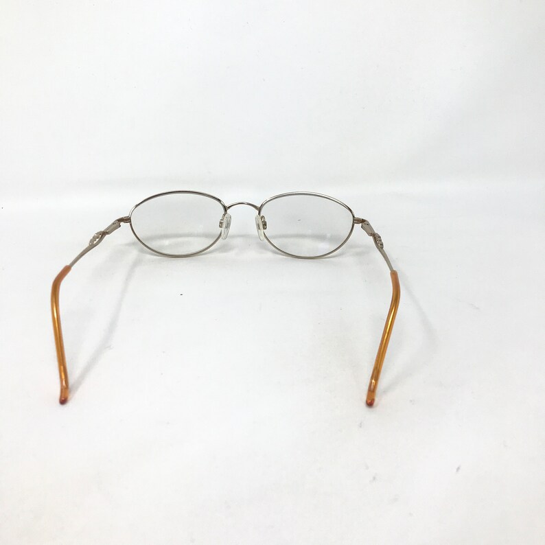 Puede incluir: Gafas de forma ovalada con montura de metal plateado y lentes transparentes. Las patillas son de color marr&oacute;n claro. Las gafas est&aacute;n sobre un fondo blanco.