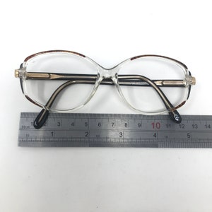 May include: Clear eyeglasses with a gold-coloured bridge and black temples. The frames have a brown tortoiseshell pattern on the upper rims. A ruler is placed below the glasses, measuring in centimetres.
