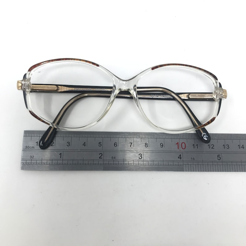 May include: Clear eyeglasses with a gold-coloured bridge and black temples. The frames have a brown tortoiseshell pattern on the upper rims. A ruler is placed below the glasses, measuring in centimetres.