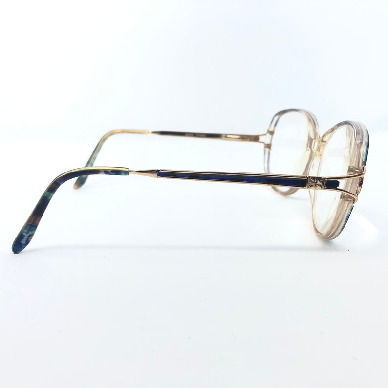 May include: Vintage eyeglasses with a gold-tone frame and clear lenses. The temples feature a blue and green marbled pattern. The frames have a geometric design at the bridge and the lenses are round.