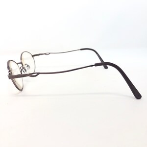 Peut inclure: Une paire de lunettes de forme ovale avec une monture en m&eacute;tal brun fonc&eacute;. Les branches sont l&eacute;g&egrave;rement incurv&eacute;es et la monture pr&eacute;sente des d&eacute;tails d&eacute;coratifs pr&egrave;s des verres. Les verres sont clairs, sur fond blanc.