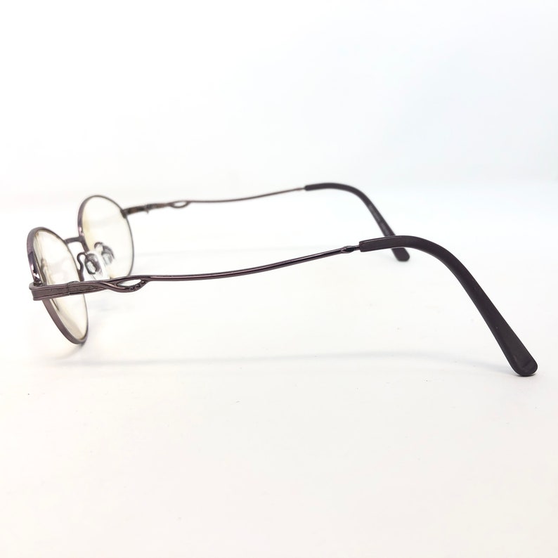 Peut inclure: Une paire de lunettes de forme ovale avec une monture en m&eacute;tal brun fonc&eacute;. Les branches sont l&eacute;g&egrave;rement incurv&eacute;es et la monture pr&eacute;sente des d&eacute;tails d&eacute;coratifs pr&egrave;s des verres. Les verres sont clairs, sur fond blanc.