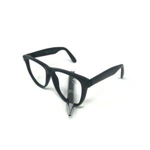 May include: Black eyeglasses with a rectangular frame and a black pen. The glasses are positioned on a white surface, with the pen resting between the lenses. The pen has a silver tip and a black body.