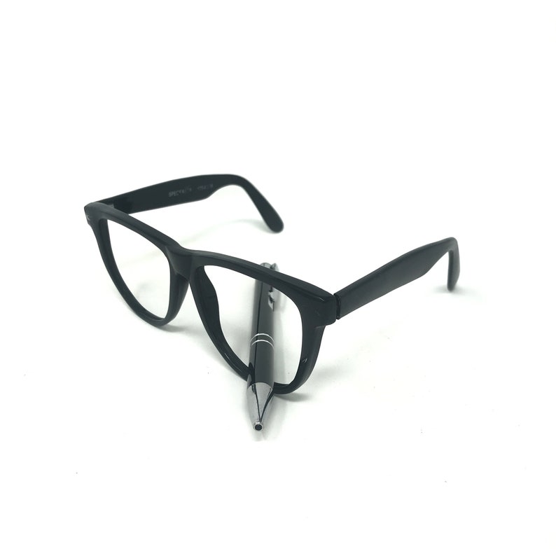 May include: Black eyeglasses with a rectangular frame and a black pen. The glasses are positioned on a white surface, with the pen resting between the lenses. The pen has a silver tip and a black body.