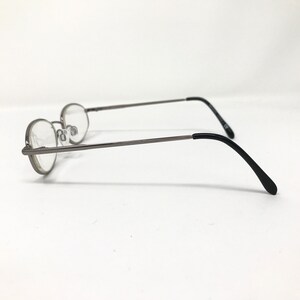 Op de afbeelding: Ovale bril met een dun, zilverkleurig metalen montuur en heldere glazen. De pootjes zijn zwart en buigen aan de uiteinden. De bril staat tegen een witte achtergrond.