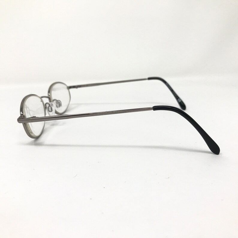 Op de afbeelding: Ovale bril met een dun, zilverkleurig metalen montuur en heldere glazen. De pootjes zijn zwart en buigen aan de uiteinden. De bril staat tegen een witte achtergrond.