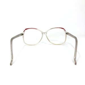 May include: Clear-framed eyeglasses with a rectangular shape. The top of the frame has a red stripe design. The arms are a light gray color. The glasses are set against a white background.