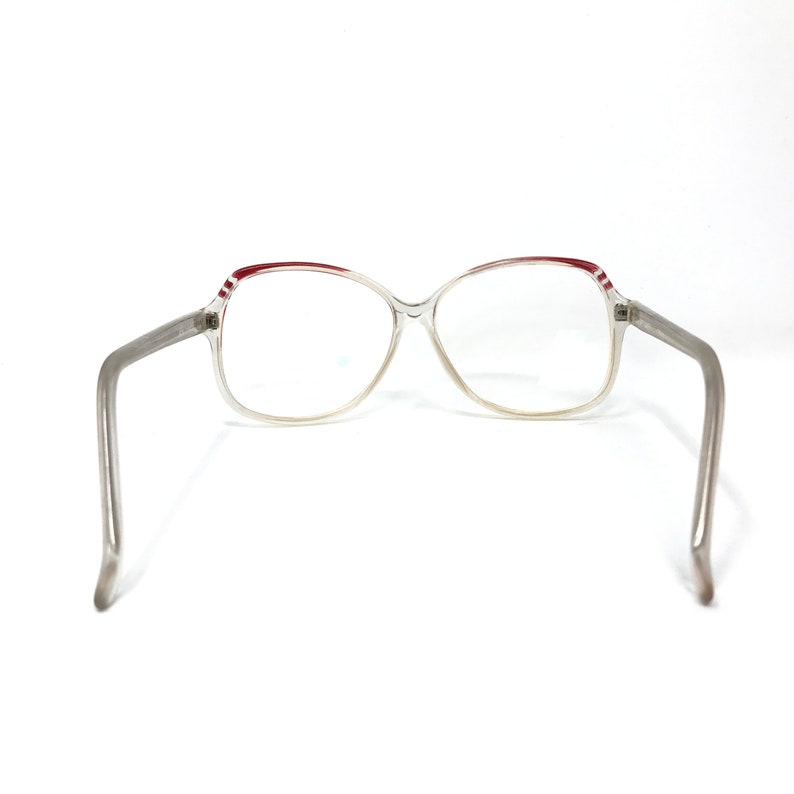 May include: Clear-framed eyeglasses with a rectangular shape. The top of the frame has a red stripe design. The arms are a light gray color. The glasses are set against a white background.