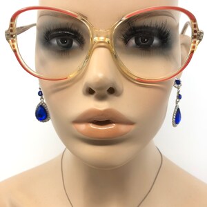 Puede incluir: Gafas de gran tama&ntilde;o con montura transparente y detalles rojos. Las gafas tienen forma rectangular y est&aacute;n sobre un maniqu&iacute;. Tambi&eacute;n se ven pendientes colgantes azules en forma de l&aacute;grima con detalles plateados.