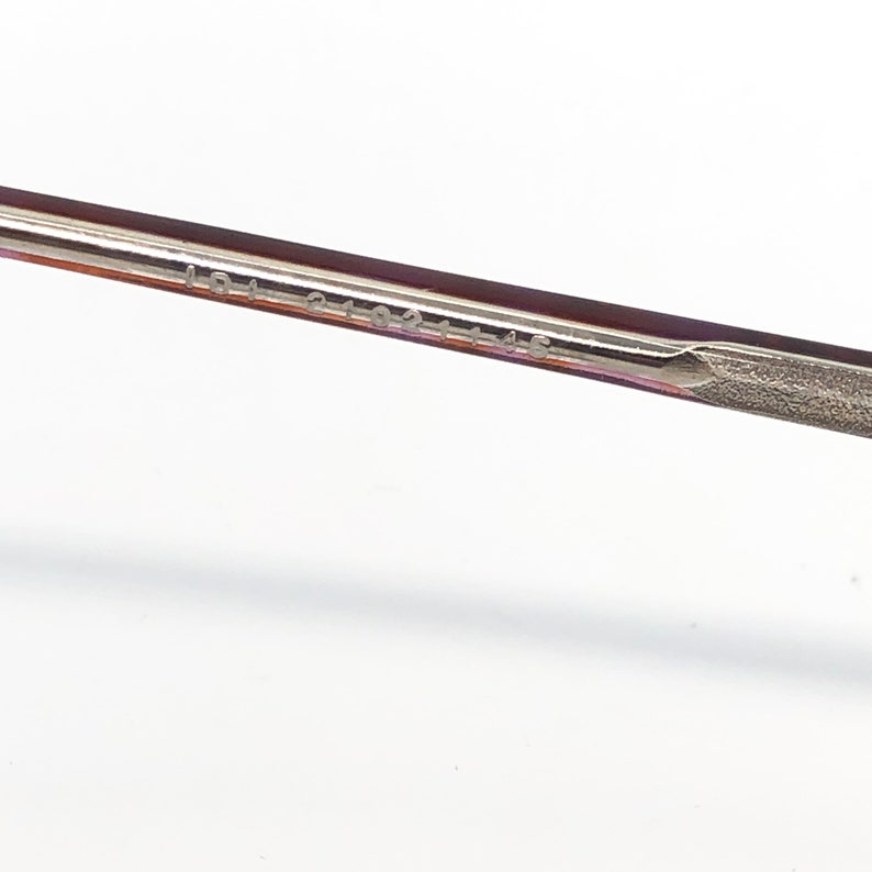 K&ouml;nnte beinhalten: Nahaufnahme eines Teils des Brillenrahmens. Der Rahmen hat eine dunkelburgunder Farbe mit einem silberfarbenen Metallb&uuml;gel. Der Text "LDJ 2102 145" ist auf dem B&uuml;gel aufgedruckt.