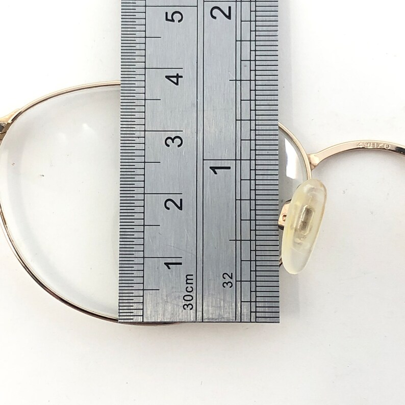 K&ouml;nnte beinhalten: Nahaufnahme einer goldgerahmten Brille mit klaren Gl&auml;sern. Ein Lineal liegt neben der Brille und zeigt Messungen in Zentimetern an. Die Brille hat ein klassisches Design mit Nasenpads.