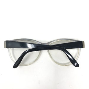 May include: Clear-framed eyeglasses with black temples and a black upper frame. The glasses have a retro style, with a slightly curved shape. The lenses are clear, and the frame has a subtle speckled design.