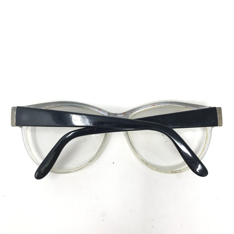 May include: Clear-framed eyeglasses with black temples and a black upper frame. The glasses have a retro style, with a slightly curved shape. The lenses are clear, and the frame has a subtle speckled design.