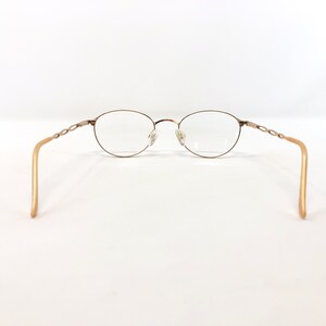 May include: Oval-shaped eyeglasses with a gold-colored metal frame and clear lenses. The temples are a light gold color and feature a decorative chain-link design near the hinges. The glasses are set against a white background.