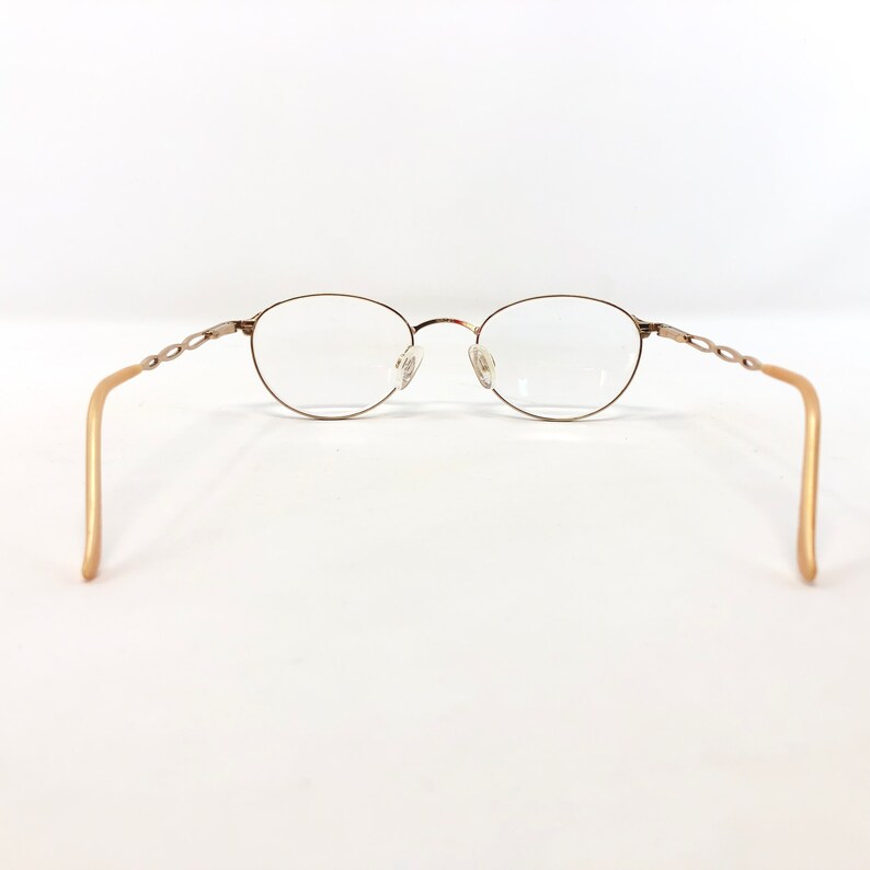 May include: Oval-shaped eyeglasses with a gold-colored metal frame and clear lenses. The temples are a light gold color and feature a decorative chain-link design near the hinges. The glasses are set against a white background.