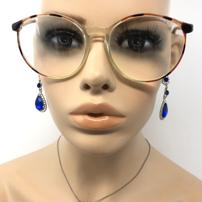 K&ouml;nnte beinhalten: Klare Brille mit Schildpattmuster an den oberen R&auml;ndern. Die Brille befindet sich auf einem Schaufensterpuppenkopf. H&auml;ngende blaue Tropfenohrringe mit silbernen Akzenten sind sichtbar.