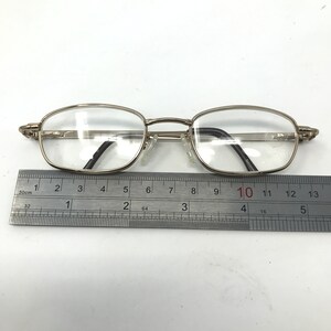 May include: Silver-framed eyeglasses with clear lenses. The glasses have an oval shape and are approximately 5 inches long. A ruler is placed beneath the glasses for measurement. The frame has a decorative design on the temples.