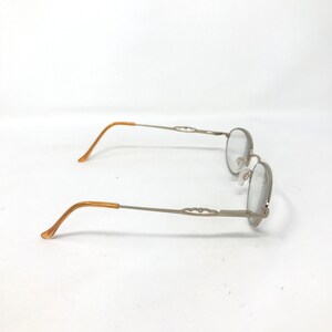 Puede incluir: Gafas plateadas con lentes ovaladas y patillas decorativas. Las patillas son de color marr&oacute;n claro. Las gafas est&aacute;n sobre un fondo blanco.