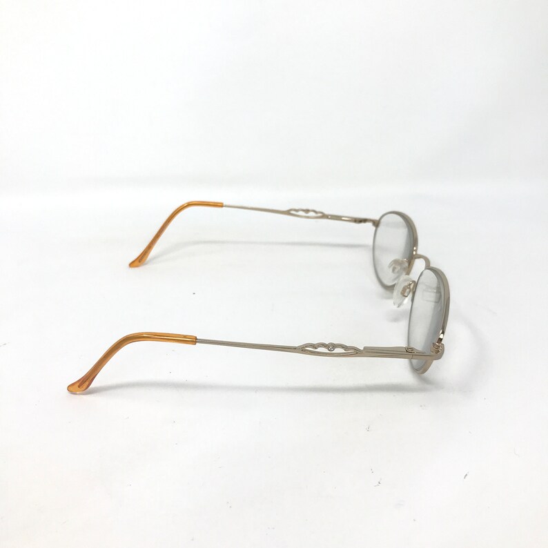 Puede incluir: Gafas plateadas con lentes ovaladas y patillas decorativas. Las patillas son de color marr&oacute;n claro. Las gafas est&aacute;n sobre un fondo blanco.