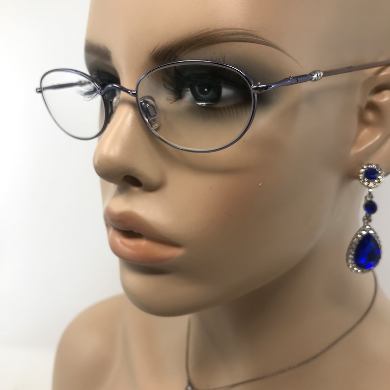 May include: A close-up of a mannequin wearing oval-shaped, light purple eyeglasses with a thin metal frame. The mannequin also wears a pair of dangling earrings with large, blue teardrop-shaped stones and a matching necklace. The background is plain white.