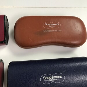 May include: A collection of eyeglass cases in various colors. A brown case with the "Specsavers Opticians" logo and website address is in the center. A dark blue case and a burgundy case are also visible.
