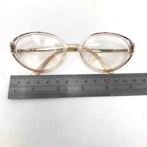 May include: Oval-shaped eyeglasses with clear lenses and a light-colored frame. The frame has a gold-colored bridge and temples. The top of the frame has a decorative pattern. A ruler is placed below the glasses, showing measurements in inches.