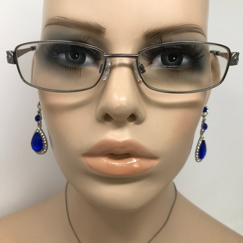 May include: Silver-framed eyeglasses with rectangular lenses are displayed on a mannequin. The glasses have a subtle design on the temples. Dangling earrings with large, blue teardrop-shaped gems and smaller blue accents are also visible.