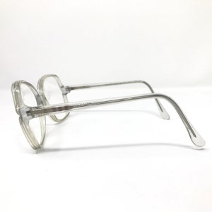 May include: Clear, octagonal-shaped eyeglasses with a transparent frame. The glasses have a classic design with a bridge and temples. The lenses are clear, and the frame is a light gray color. The glasses are set against a white background.