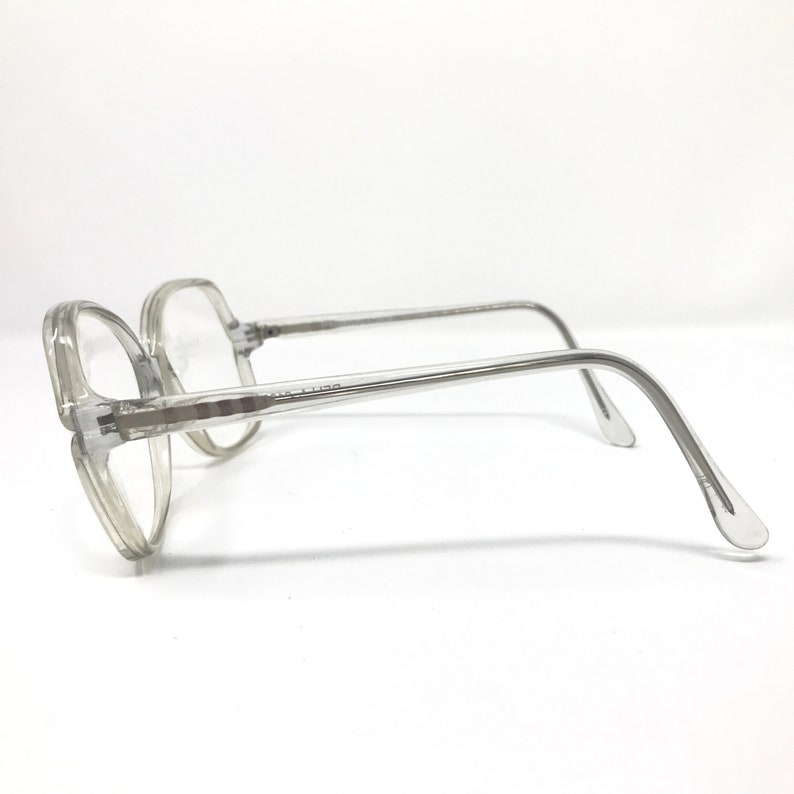 May include: Clear, octagonal-shaped eyeglasses with a transparent frame. The glasses have a classic design with a bridge and temples. The lenses are clear, and the frame is a light gray color. The glasses are set against a white background.