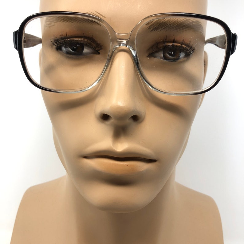 K&ouml;nnte beinhalten: Brille mit transparentem Rahmen und schwarzen Akzenten. Die Brille hat eine quadratische Form und wird auf einem Schaufensterkopf pr&auml;sentiert. Die Rahmen sind dick und die Gl&auml;ser klar. Der Stil ist Vintage.