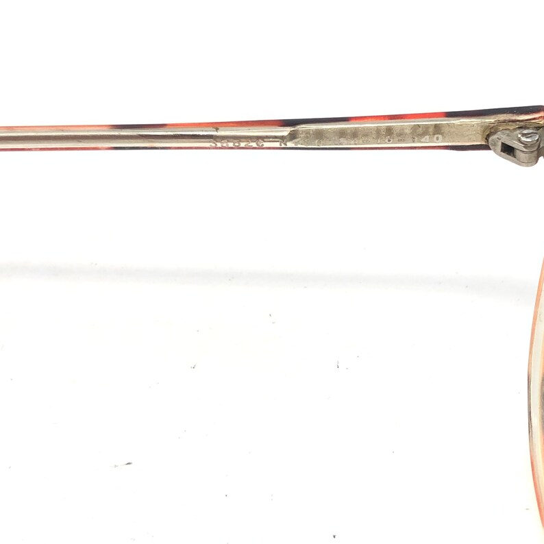 K&ouml;nnte beinhalten: Nahaufnahme des B&uuml;gels einer Brille. Der B&uuml;gel weist eine Mischung aus roten, braunen und schwarzen Mustern auf. Das Metallscharnier ist sichtbar, und der Text "58020 N" und "140" ist auf dem B&uuml;gel aufgedruckt.