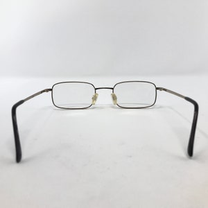 May include: Rectangular eyeglasses with a thin, dark metal frame and clear lenses. The temples are a lighter metal color, and the earpieces are black. The glasses are set against a white background.