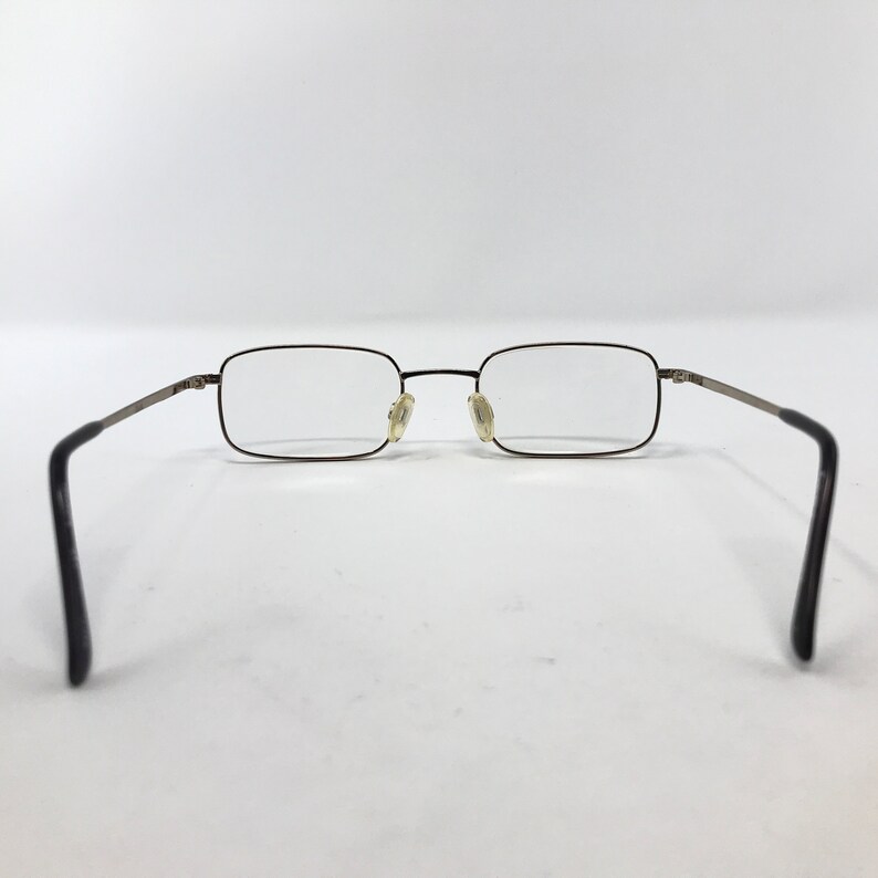 May include: Rectangular eyeglasses with a thin, dark metal frame and clear lenses. The temples are a lighter metal color, and the earpieces are black. The glasses are set against a white background.