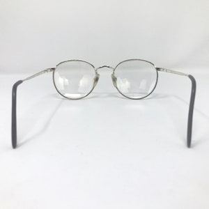 Peut inclure: Lunettes &agrave; monture argent&eacute;e avec verres ronds. Les lunettes ont un design classique avec une monture en m&eacute;tal fin et des branches gris fonc&eacute;. Les verres sont clairs, sur fond blanc.