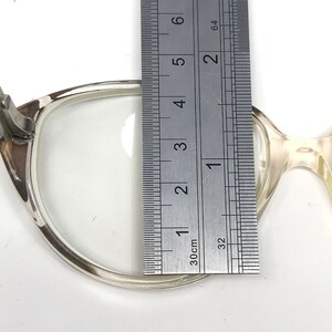 May include: Close-up of a pair of eyeglasses with a ruler measuring the lens. The eyeglasses have a light-colored frame. The ruler shows a measurement of approximately 1.5 inches.