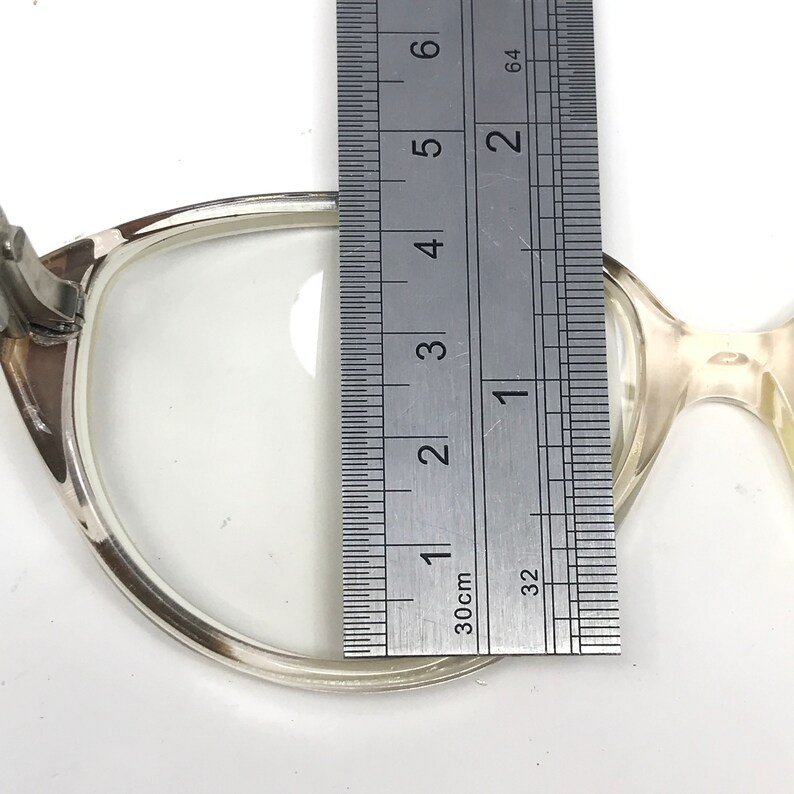 May include: Close-up of a pair of eyeglasses with a ruler measuring the lens. The eyeglasses have a light-colored frame. The ruler shows a measurement of approximately 1.5 inches.
