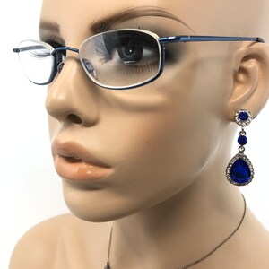 May include: A mannequin head wearing blue-rimmed eyeglasses and dangling earrings. The earrings feature a cluster of small, clear stones around a large, teardrop-shaped, deep blue stone. The glasses have a thin, blue frame.