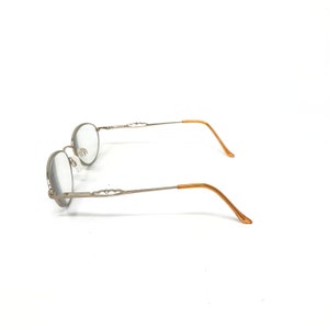 Puede incluir: Gafas de forma ovalada con montura plateada y patillas doradas. Vista lateral, las lentes son claras. Las patillas presentan detalles decorativos. El conjunto se presenta sobre un fondo blanco.
