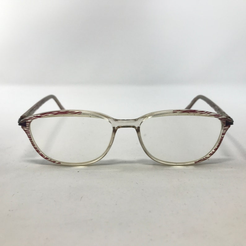 May include: Clear eyeglasses with a thin, oval frame. The rims feature a red and white striped pattern. The temples are a dark brown color. The glasses are set against a white background.